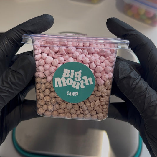 Close-up image of a clear plastic tub of candy from Big Mouth Candy, held by gloved hands. The tub is filled with two distinct layers of small