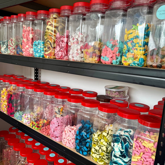 Shelves filled with clear plastic jars of colourful sweets, each topped with a red lid. The jars contain a wide variety of pick-and-mix treats.