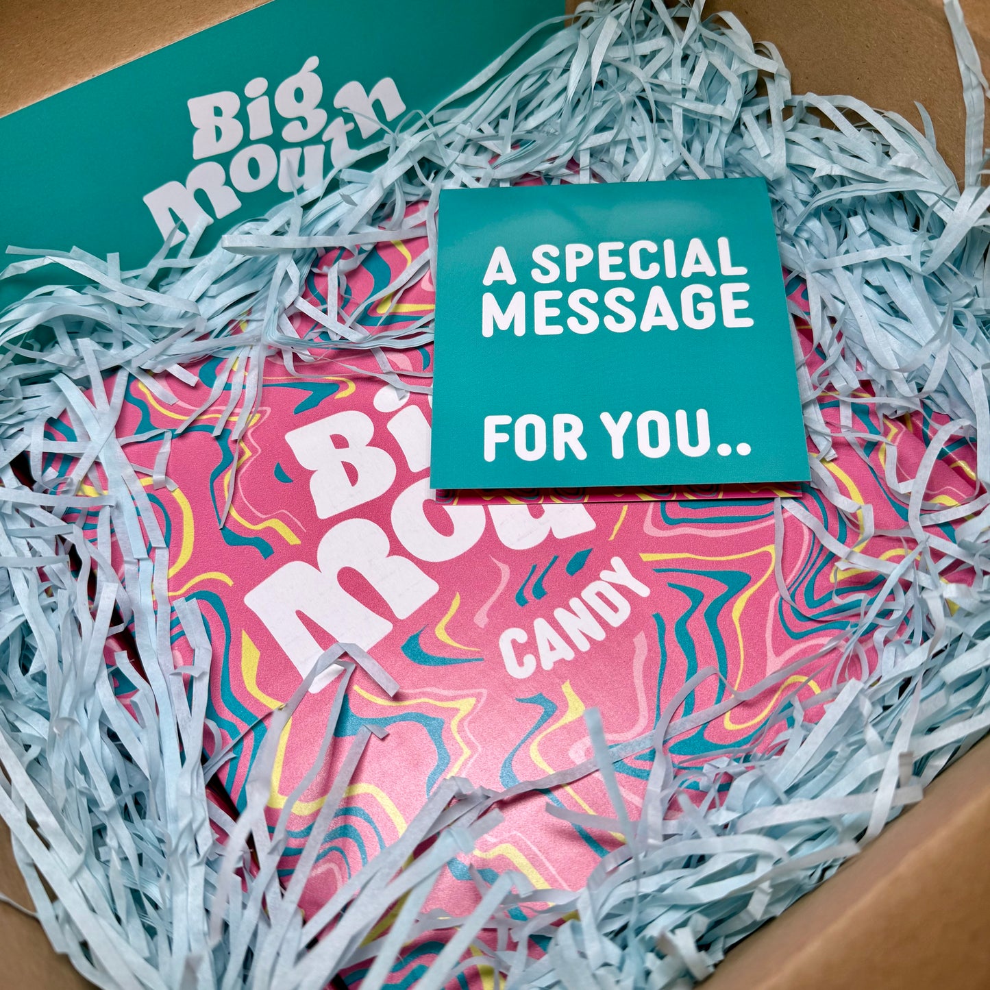 Big Mouth Candy gift box surrounded by light blue shredded tissue paper, featuring a teal card that reads “A special message for you.” Perfect for gifting and sweet surprises.