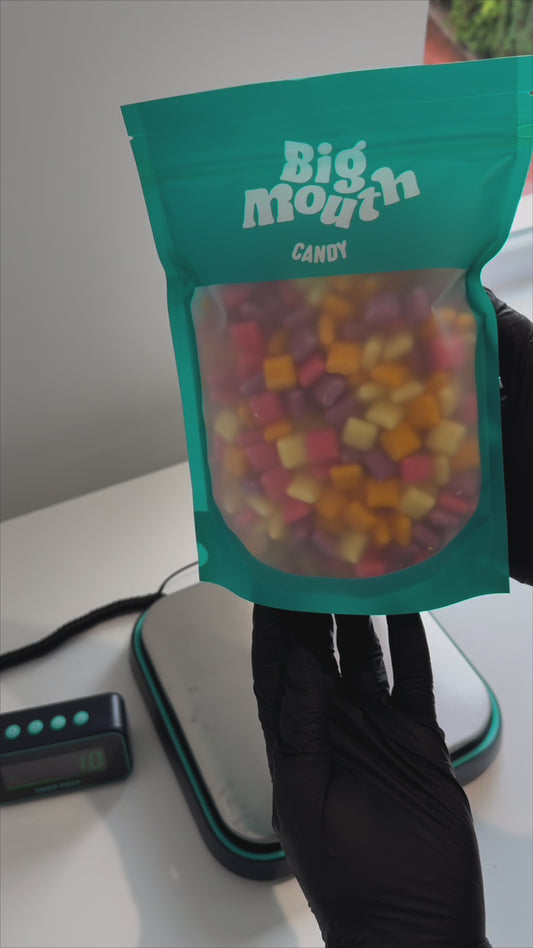 Person holding a resealable teal pouch of Big Mouth Candy, filled with colorful square-shaped sweets. The packaging features a frosted window showcasing the candy inside. Perfect for pick and mix uklovers looking for a sweet treat