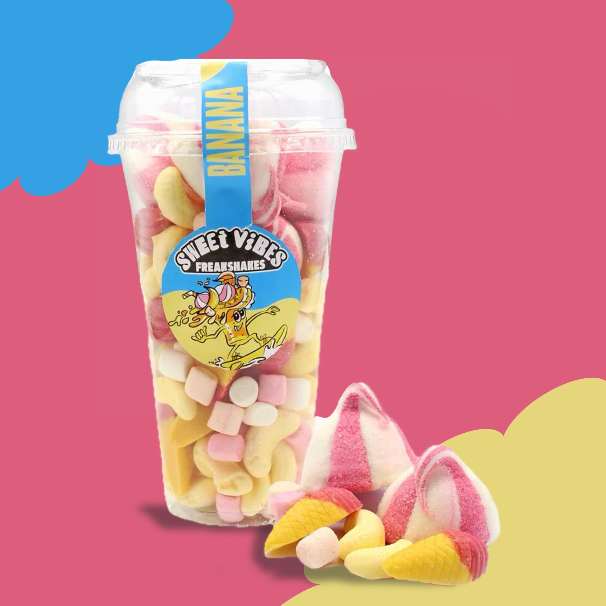 A colourful plastic cup filled with banana-themed pick-and-mix sweets, including foam bananas, mini marshmallows, and pink-and-yellow gummy cones. The cup is labeled “Sweet Vibes Freakshakes Banana” with a playful cartoon design, and is placed on a vibrant pink and yellow background with a few sweets displayed beside it.
