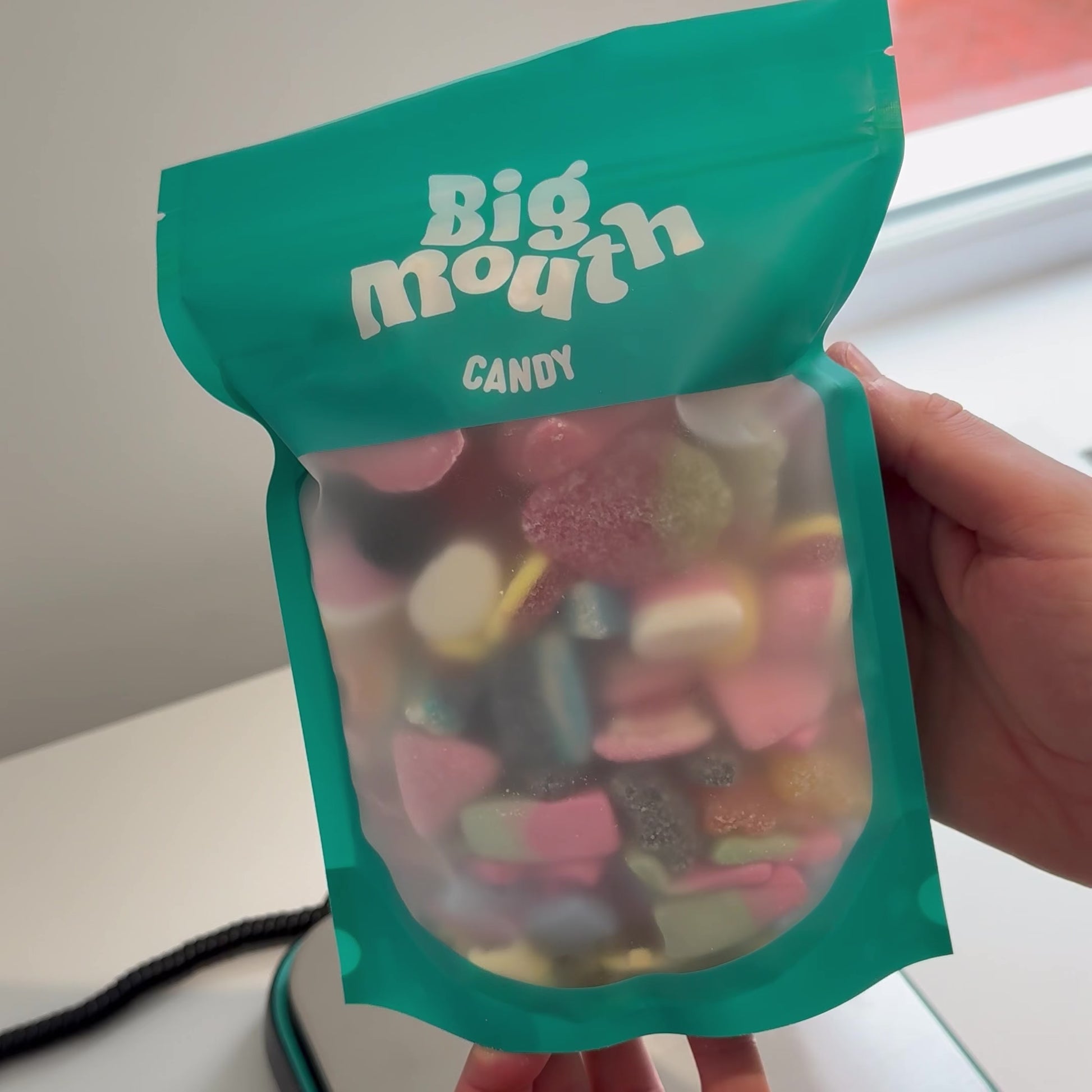 Person holding a resealable teal pouch of Big Mouth Candy, filled with colorful square-shaped sweets. The packaging features a frosted window showcasing the candy inside. Perfect for pick-and-mix lovers looking for a sweet treat
